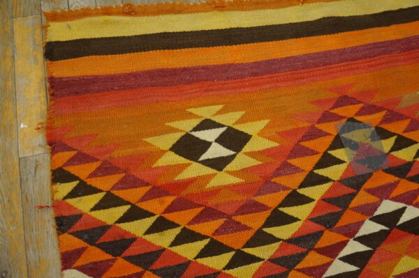 Geometric flat-weave rug featuring orange, red, yellow, and brown colors lying on worn wooden floors.