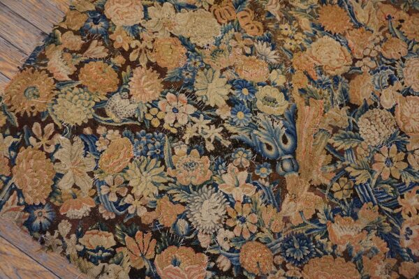 Worn floral rug fragment in tan, blue, and peach with visible stitching rests on brown wood planks.