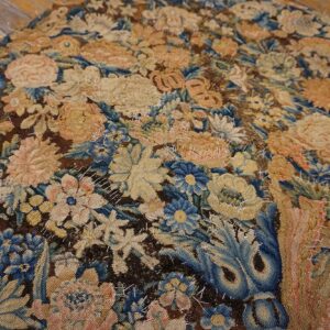 Low-pile rug with dense floral patterns in muted blue, tan, and dark brown, accented by rough white stitching.