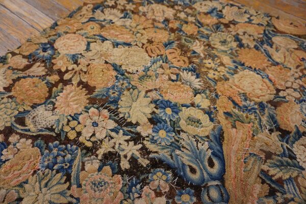Low-pile rug with dense floral patterns in muted blue, tan, and dark brown, accented by rough white stitching.