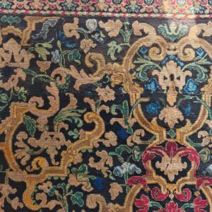 Low-pile rug with dark ground, featuring dense scrolling florals in gold, teal, red, and blue.