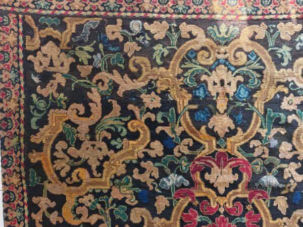 Low-pile rug with dark ground, featuring dense scrolling florals in gold, teal, red, and blue.