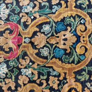 Detailed low-pile rug close-up showing gold, magenta, blue, and green scrolling botanical motifs on a dark ground.