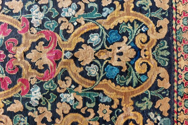 Detailed low-pile rug close-up showing gold, magenta, blue, and green scrolling botanical motifs on a dark ground.