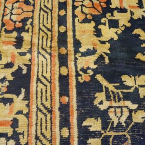 Detailed view of a navy rug featuring tan and coral traditional motifs and geometric borders next to wood flooring.