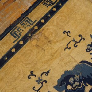 Worn yellow rug with dark blue mythological figures and geometric border on distressed wood floor.