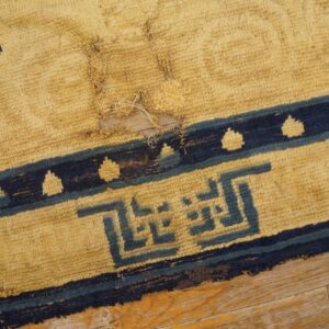Close-up of a damaged yellow high-pile rug with dark blue geometric patterns lying on wood flooring.