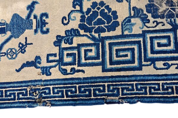 Detailed close-up of a cream rug featuring deep blue fretwork and stylized floral patterns near the worn edge.