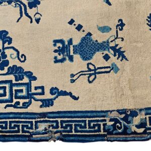 Detail of a low-pile rug showing blue floral, ribbon, and key patterns on a cream background.