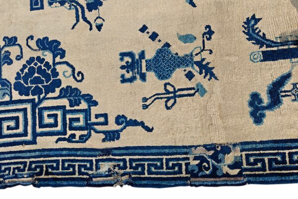 Detail of a low-pile rug showing blue floral, ribbon, and key patterns on a cream background.