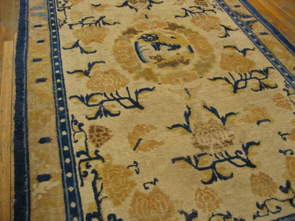 Tan rug with bold navy blue and golden floral patterns, centered motifs, and striped blue borders on wood.
