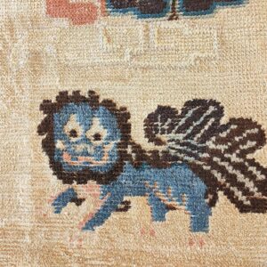 Textured rug close-up with a blue and brown winged creature pattern on a tan background.