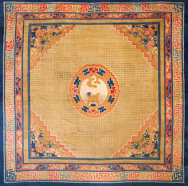 Square tan rug with a central white bird medallion, surrounded by complex blue and red geometric borders.