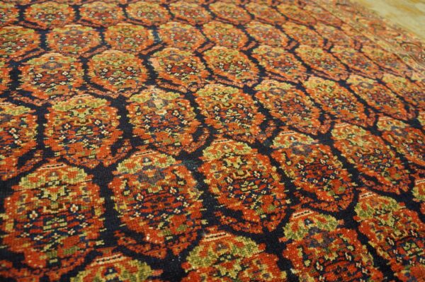 Angled view of a low-pile rug featuring a dense lattice pattern of red-orange, navy, and green medallions.