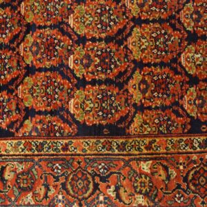 Traditional red, navy, and gold rug showing complex patterns and a detailed border against light wood planks.