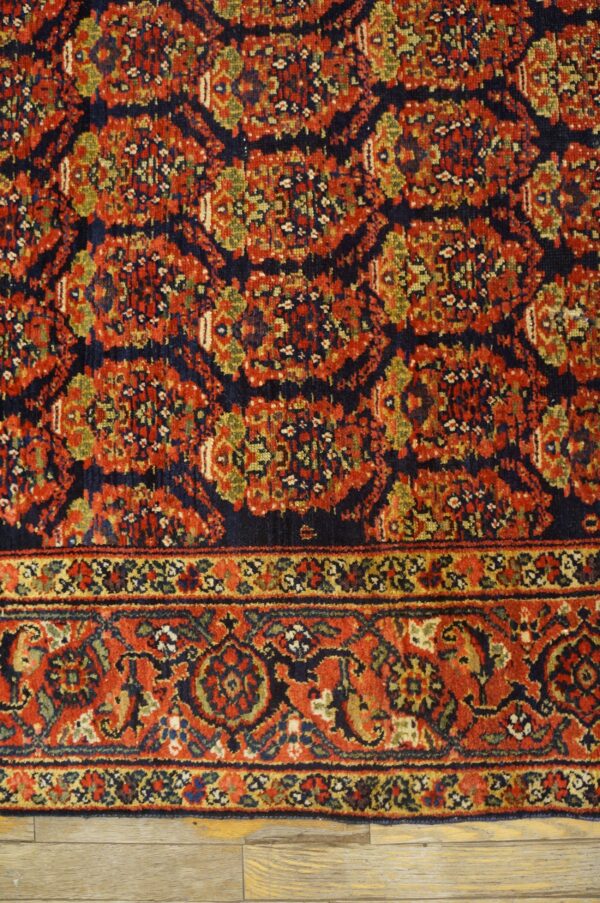 Traditional red, navy, and gold rug showing complex patterns and a detailed border against light wood planks.