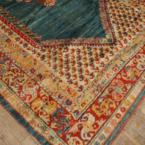 Textured rug corner featuring a teal field and thick patterned borders in red, cream, and yellow on wood planks.