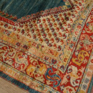A high-pile rug with red, cream, and teal traditional borders is placed on weathered wood flooring.