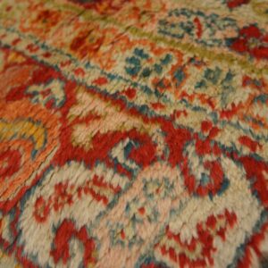High-pile rug texture closeup featuring a detailed pattern in red, orange, cream, and blue-green.