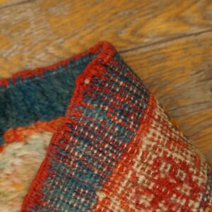 Folded rug edge displaying coarse red, blue, and cream geometric weave against wood flooring.