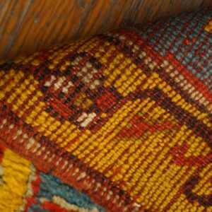 Close-up of a folded geometric rug in yellow, red, and teal, resting on rustic wood flooring.