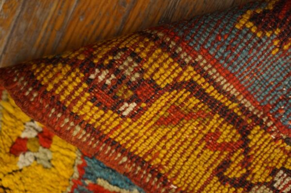 Close-up of a folded geometric rug in yellow, red, and teal, resting on rustic wood flooring.