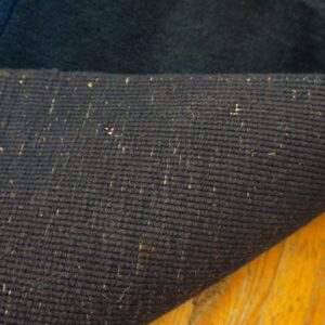 Close-up of a dark blue rug's ribbed backing with light flecks, curled over glossy yellow wood.