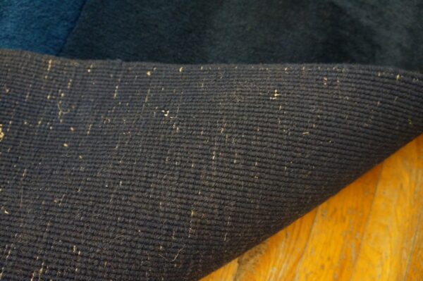 Close-up of a dark blue rug's ribbed backing with light flecks, curled over glossy yellow wood.