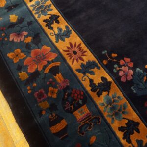 Ornate floral border in rust, teal, and gold on a dark navy rug lying on yellow wood planks.