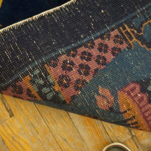 The back of a rug reveals tight weaving in navy, rose, and teal over yellow-toned wood flooring.