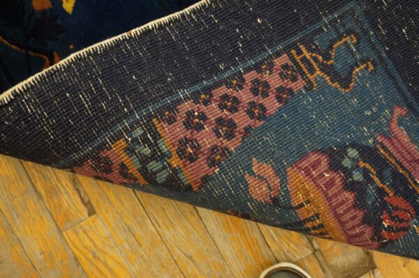 The back of a rug reveals tight weaving in navy, rose, and teal over yellow-toned wood flooring.