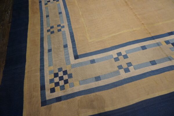 Geometric flatweave rug corner showing a tan field and stepped borders in navy, blue, and light blue.