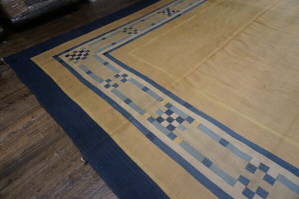 Low-pile tan rug with a bold geometric blue border on dark wood flooring.