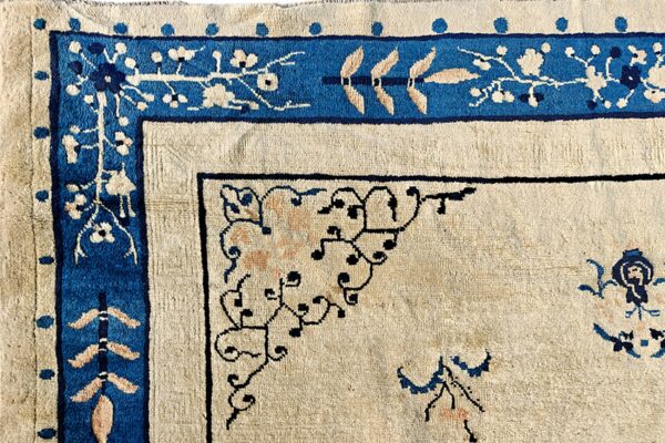 Cream rug corner featuring a thick blue border with ivory floral motifs and fine black scrollwork.