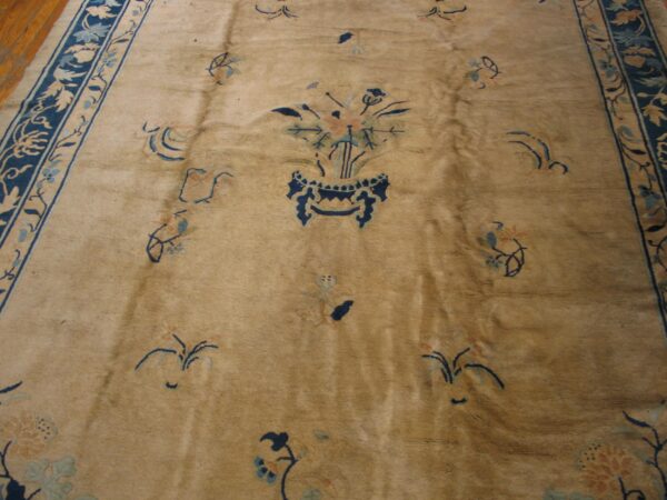 Tan low-pile rug with a blue floral vase center and wide dark blue vine border on a wood floor.