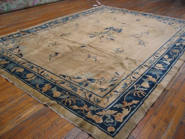 Tan area rug featuring a thick blue floral border and scattered blue designs sits on distressed wood flooring.