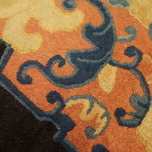 Thick pile rug detail featuring scrolling dark blue and gold motifs on a rust background near a black edge.