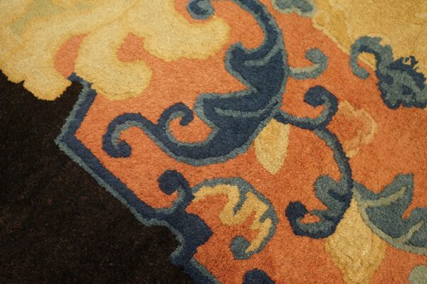 Thick pile rug detail featuring scrolling dark blue and gold motifs on a rust background near a black edge.