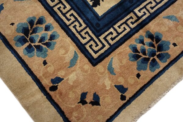 Tan field rug corner displaying blue floral clusters and a greek key border in dark blue and beige.