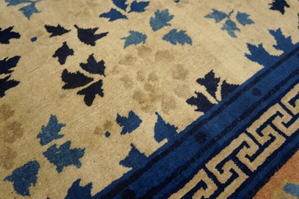 Tan rug with scattered dark and medium blue leaf patterns and a detailed greek key border.