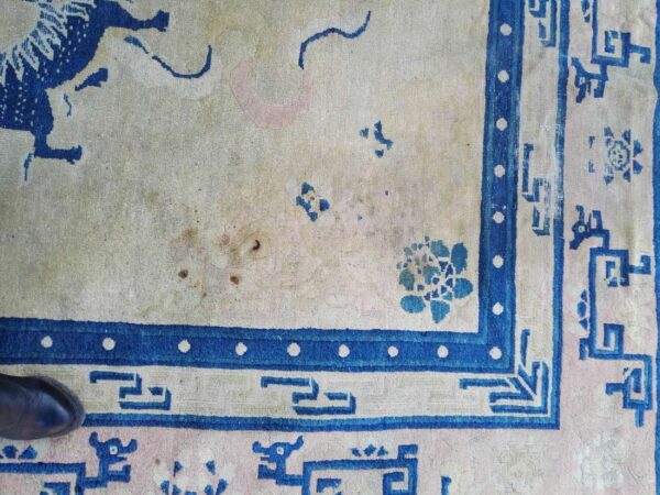 Worn cream rug with dark blue animal and floral motifs, geometric key borders, and visible staining.