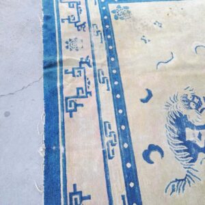 Worn beige and cobalt blue rug featuring geometric and animal patterns displayed on cracked gray concrete.