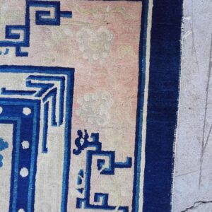 Detail of a rug showing a pink field with cloud motifs and a blue geometric fretwork border on concrete.