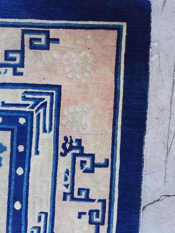 Detail of a rug showing a pink field with cloud motifs and a blue geometric fretwork border on concrete.