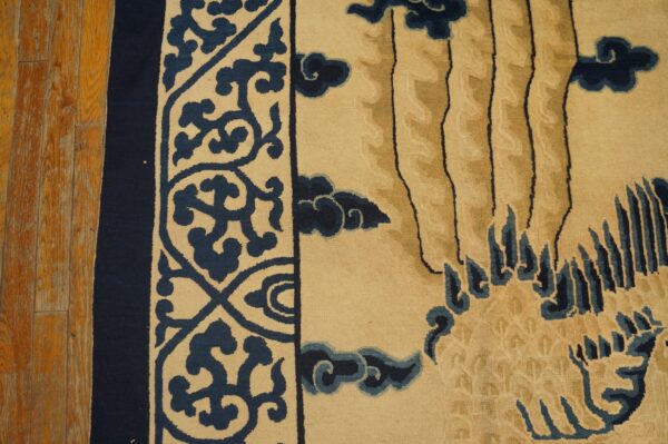 Low-pile rug in beige and navy blue with scrollwork border and stylized nature motifs rests on distressed wood flooring.