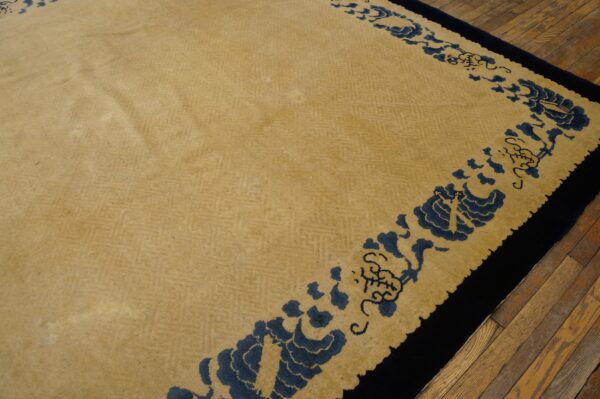 Tan rug with subtle herringbone field pattern and navy blue scrolling border on wood planks.