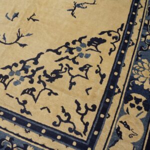 Beige rug corner featuring complex dark blue floral and scrollwork designs, resting on distressed hardwood flooring.