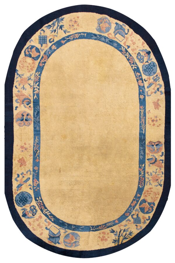 An oval tan rug featuring a decorative border with blue, peach, and navy traditional motifs.