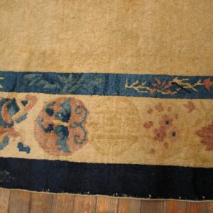 Closeup of a tan rug with patterned blue and mauve borders ending in a solid dark navy band on wood flooring.