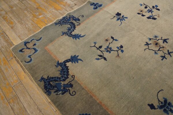 Muted blue-gray rug featuring a dark blue dragon border and florals lying on distressed wooden flooring.